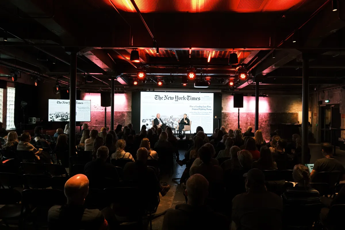 Audience in a dark hall in front of a stage with a panel and a big screen showing The New York Times