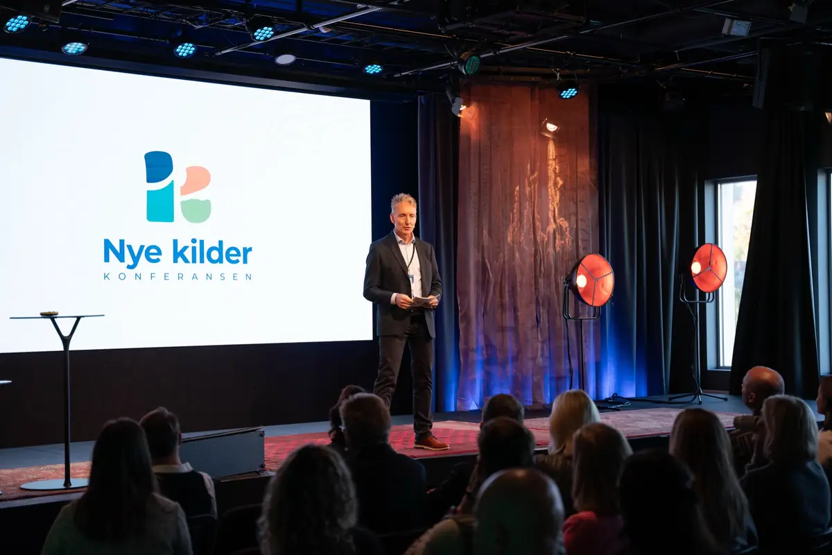 Host on stage before a large screen with the Nye Kilder logo and the audience in the hall