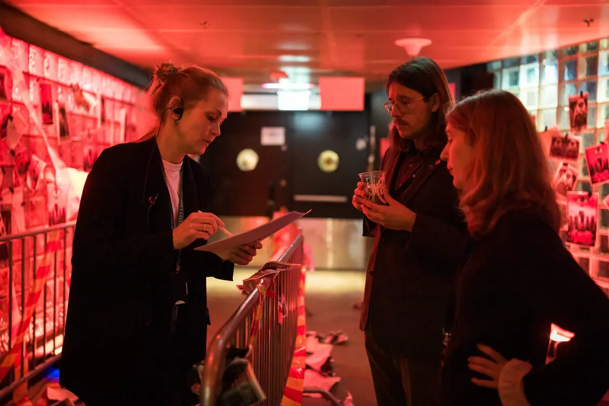 Three people in a red-lit corridor with posters and barriers at Kvarteret