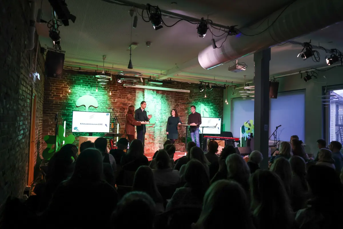Panel debate on stage with green light and brick walls, seen from behind the audience