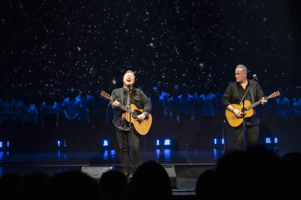 Two guitarists on stage in front of a star curtain with the youth choir in blue light behind