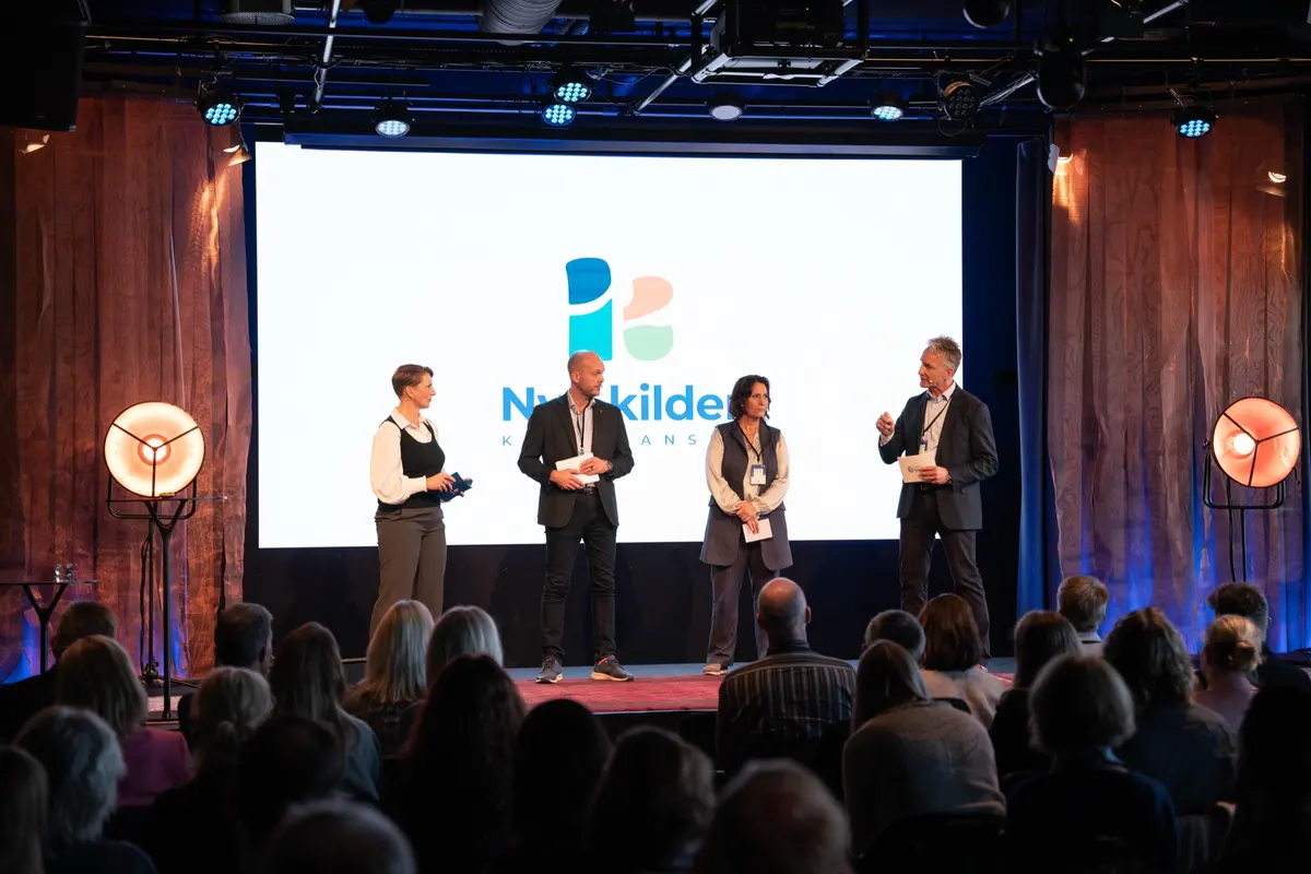 Four people standing on stage in front of the Nye kilder screen while the audience looks on