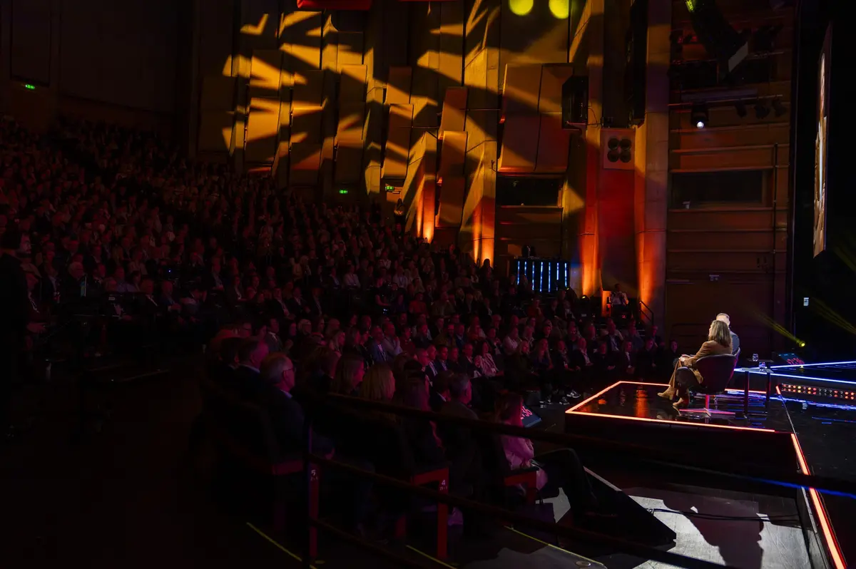 Packed hall seen from the balcony, with a person on a brightly lit stage