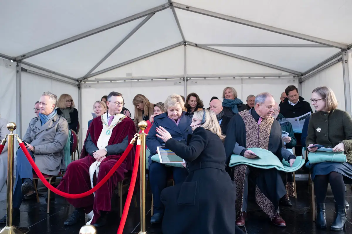 Guests in academic gowns seated in a tent behind red rope barriers