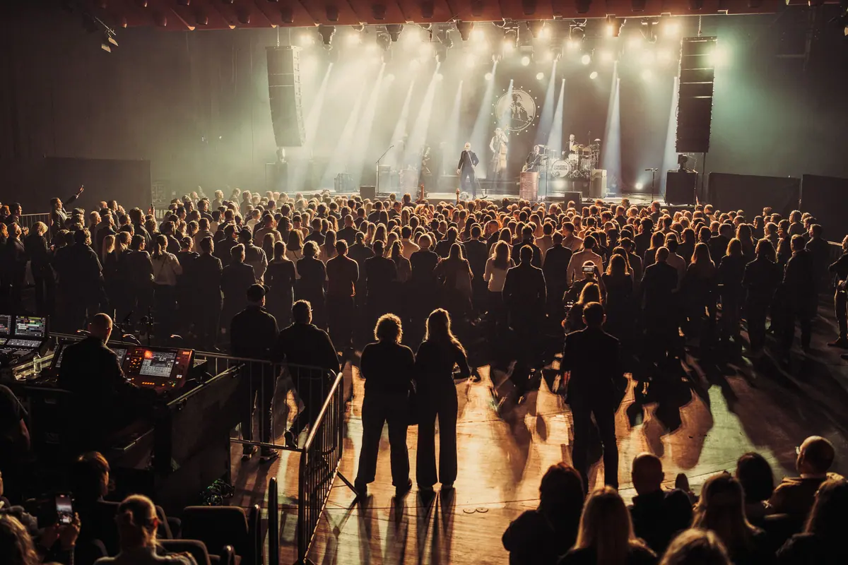 Concert audience seen from behind facing a stage with lighting rig and haze