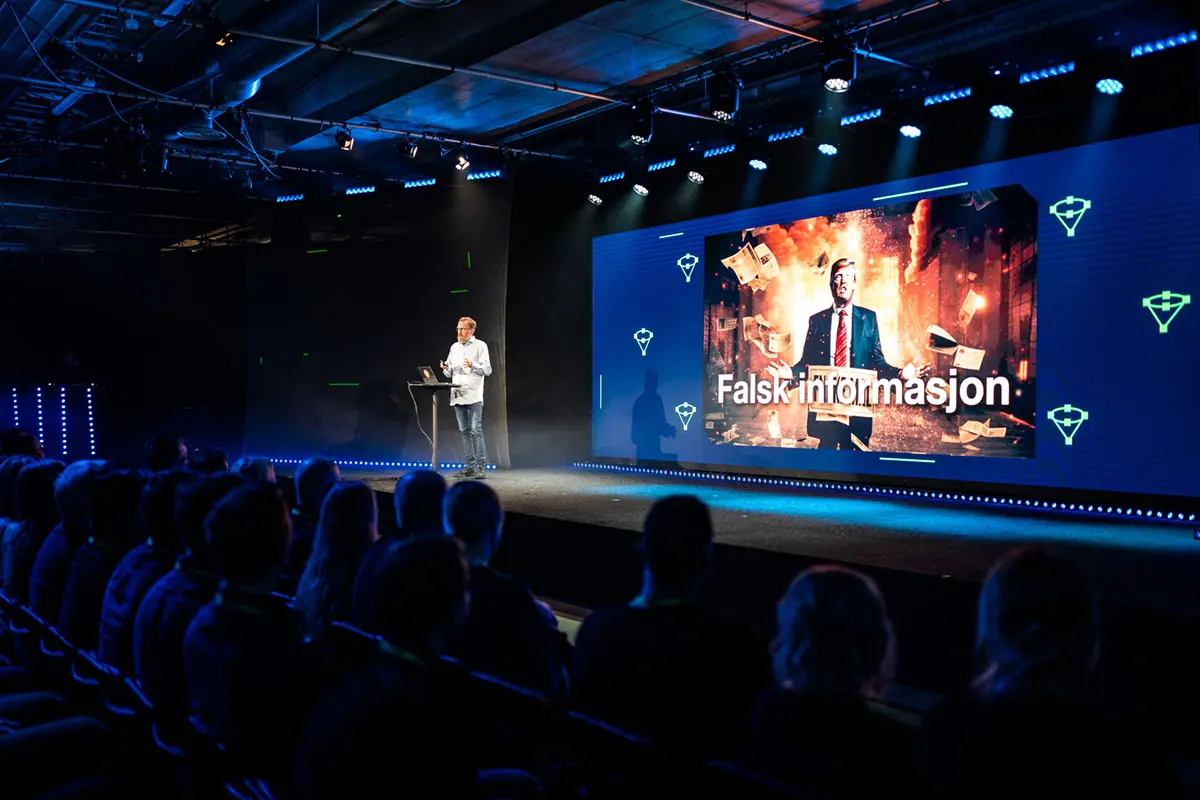 Speaker on the Volum stage before a large screen reading «Falsk informasjon»