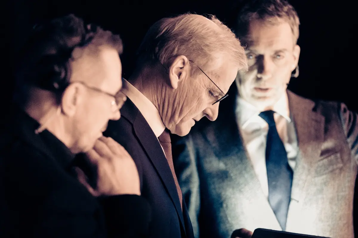 Three men in suits looking down at a screen in the backstage darkness