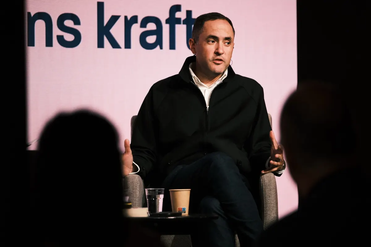 Man in a dark jacket seated on stage in front of a screen reading «Fortellingens kraft»
